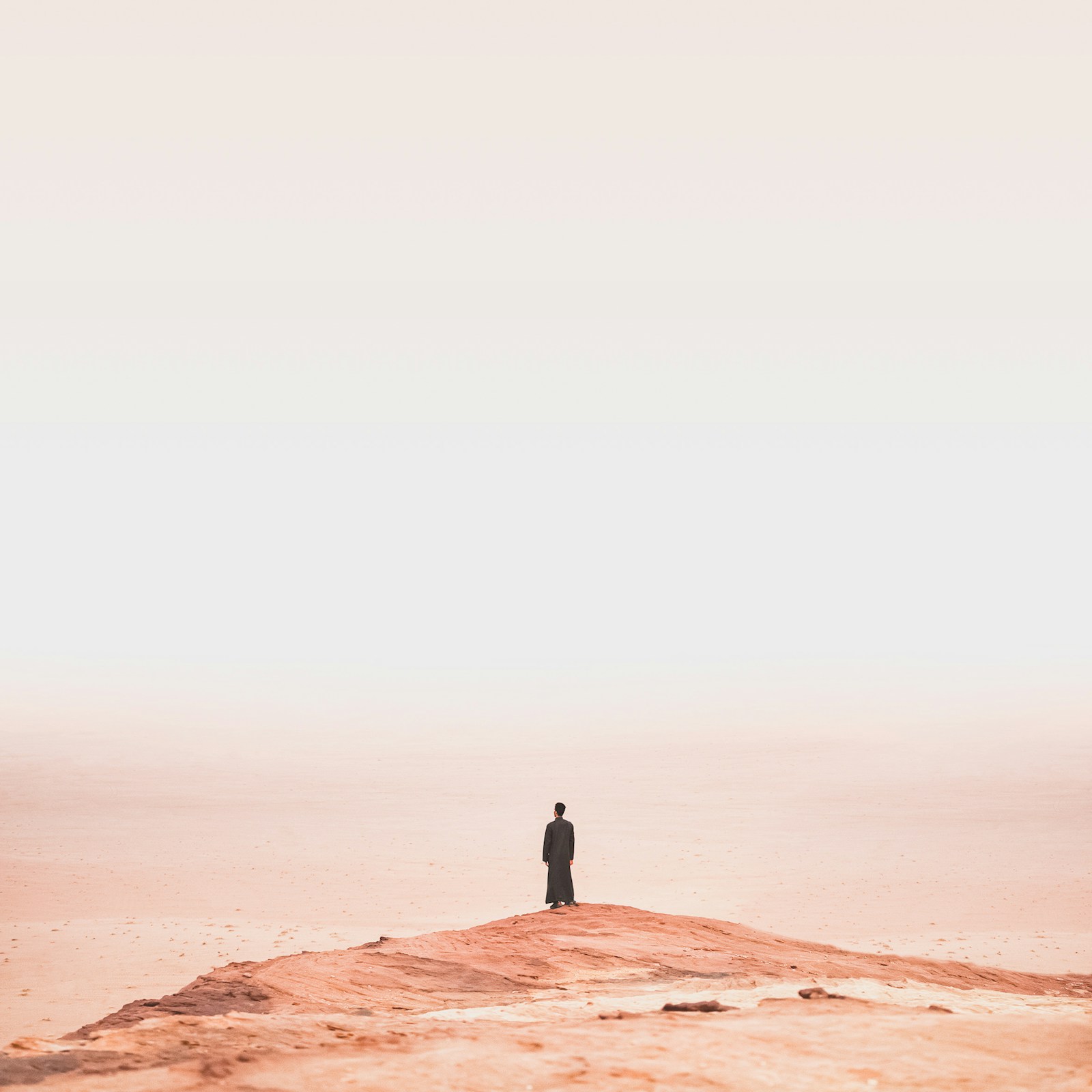 A lone robed figure standing atop a desert dune in hazy pink light