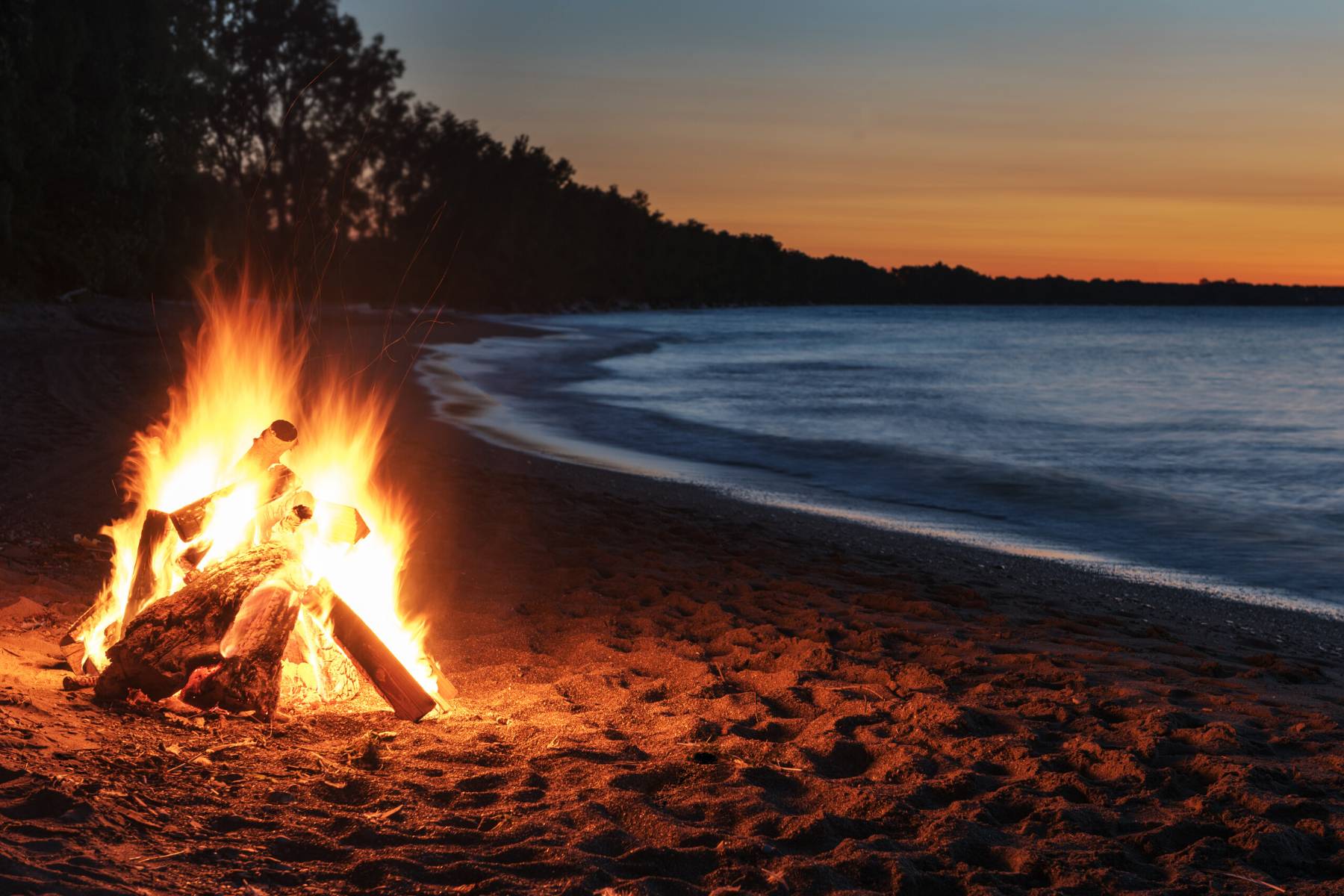 A bonfire burning on the beach at dusk, ocean in the background.