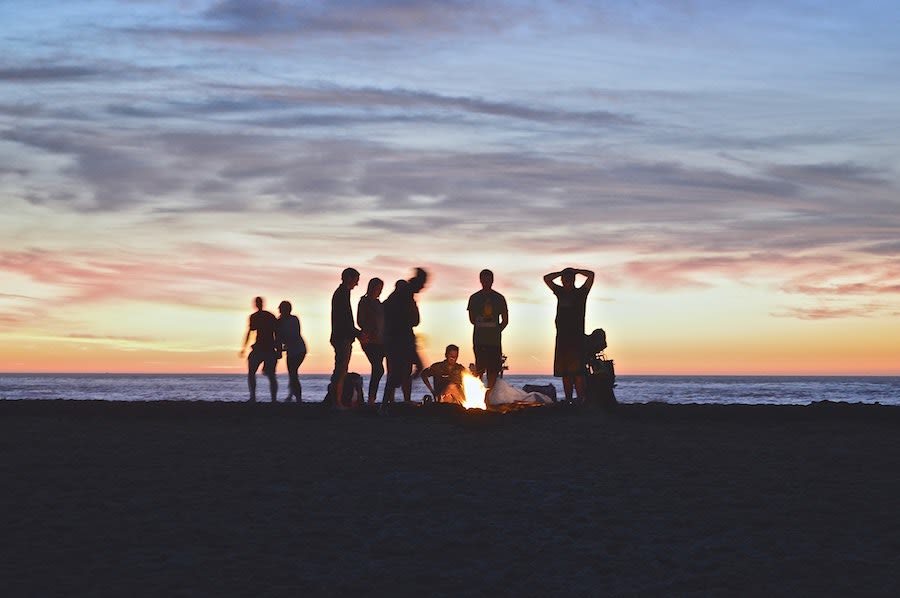 A group silhouetted around a bonfire on the beach at sunset.