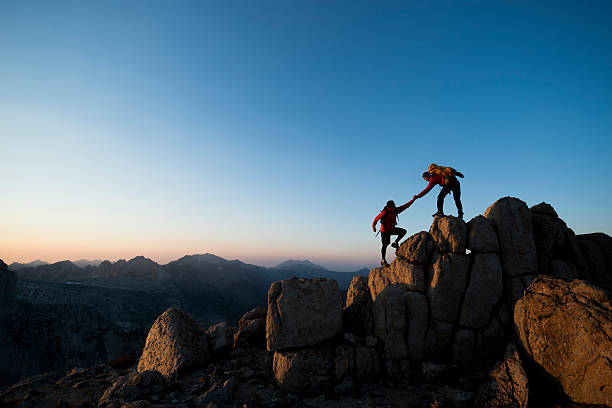 One climber reaching down to help another up a rocky summit.